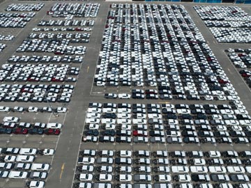 Vogelperspektive auf einen großen Parkplatz, der mit Reihen geparkter Autos gefüllt ist, überwiegend weiße Fahrzeuge, mit einigen schwarzen und roten Fahrzeugen dazwischen.