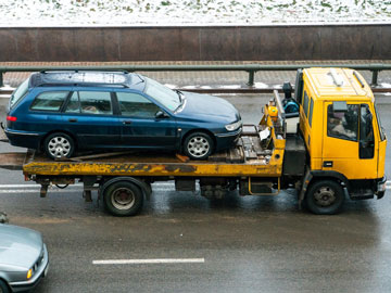 Ein blauer Wagen wird auf einem gelben Abschleppwagen auf einer nassen Straße transportiert, mit einer verschneiten Landschaft im Hintergrund.