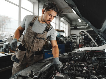 Ein Mechaniker in Arbeitsoveralls inspiziert einen Motor in einer gut beleuchteten Garage, die mit Fahrzeugen und Werkzeugen gefüllt ist.