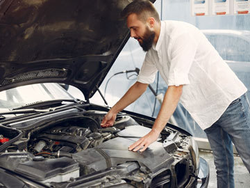 Eine Person in einem weißen Hemd untersucht den Motor eines Autos mit angehobener Motorhaube, umgeben von einer Werkstattumgebung.